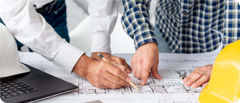 Dos profesionales revisan planos de construcción sobre una mesa, junto a un casco de seguridad y un computador portátil.