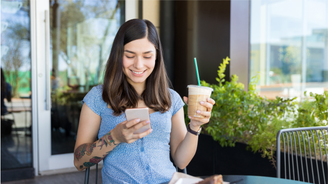 Mujer joven revisando propiedades desde su celular mientras toma un café en una terraza