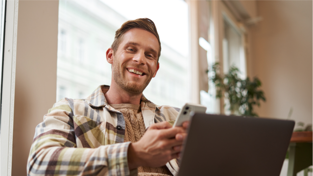 Hombre sonriente usando su celular y notebook para simular su crédito hipotecario